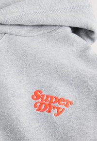 Hellgrauer Stoff-Hoodie mit gefalteter Kapuze und rotem besticktem Schriftzug "Super Dry" auf der Brust.