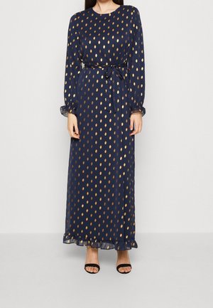 Femme portant une robe maxi bleu marine à manches longues avec des motifs goutte d'or, une ceinture nouée à la taille et des talons ouverts noirs.