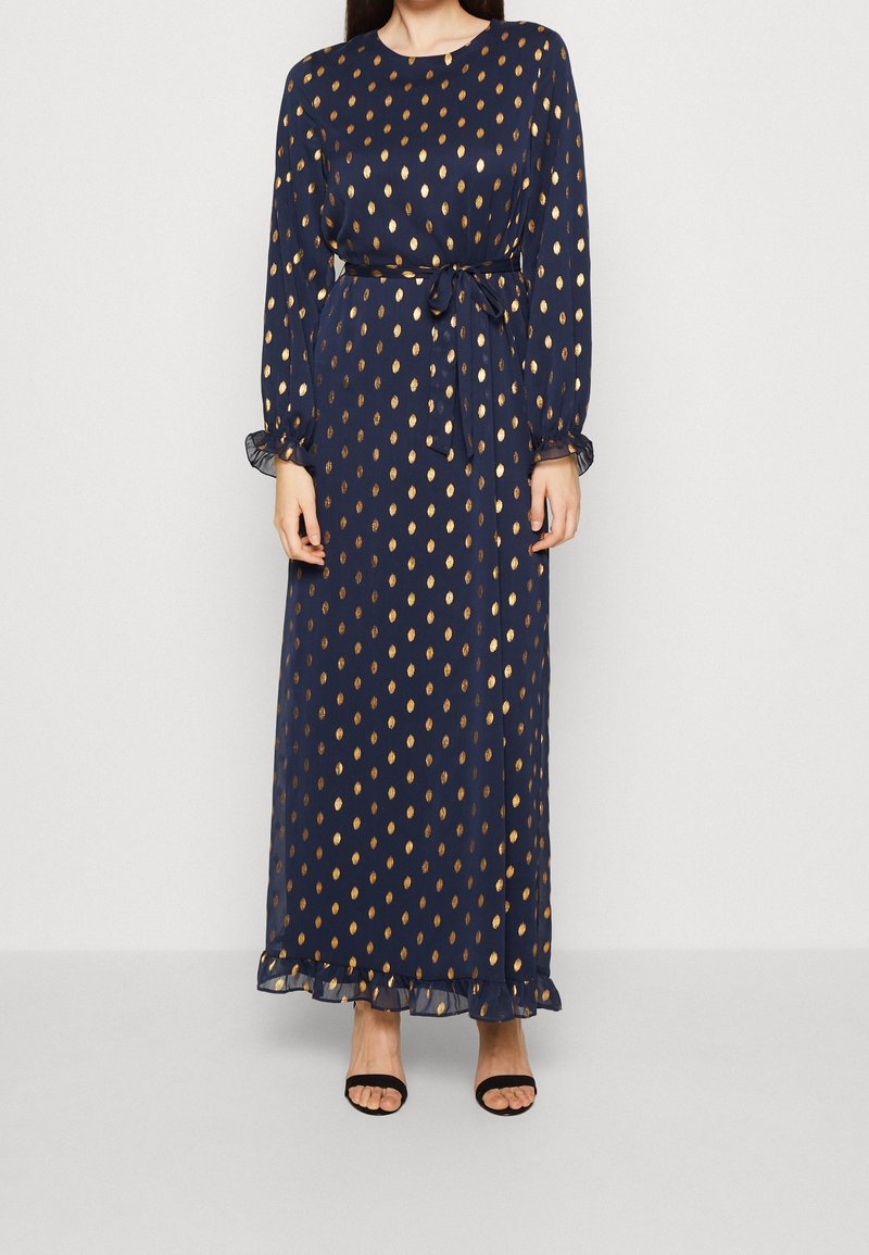 Femme portant une robe maxi bleu marine à manches longues avec des motifs goutte d'or, une ceinture nouée à la taille et des talons ouverts noirs.