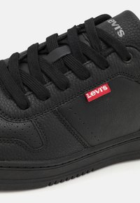 Zapatilla negra de cuero con una superficie texturizada, cordones redondos y una etiqueta roja de Levi's en el costado. Suela de goma con un patrón de banda de rodadura visible.