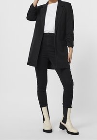 Musta bleiseri valkoisen t-paidan päällä, yhdistettynä mustiin slim-fit -farkkuihin ja kermaisiin nilkkabootseihin, joissa on paksut mustat pohjat ja sivupaneelit.