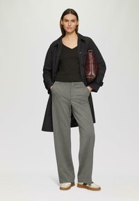 Pantalons gris longs avec poches avant, associés à un t-shirt noir à col en V et un trench-coat sombre, tenant un sac à main matelassé bordeaux.