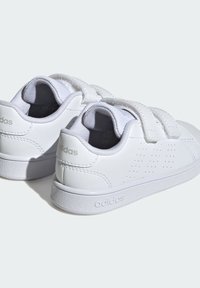 Para białych sneakersów Adidas z rzepami, teksturowanymi podeszwami oraz markowymi detalami na piętach i śródstopiu, pokazane z tyłu pod kątem.