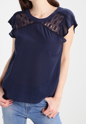 Kobieta nosząca granatową bluzkę z koronkowymi detalami na górnej części klatki piersiowej oraz falbaniaste rękawy, w połączeniu z jasnoniebieskimi dżinsami.