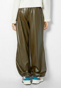 Olivgröna byxor i konstläder med avslappnad passform, elastisk midja och dragkedjeslutningar, kombinerat med vita och blå sneakers.
