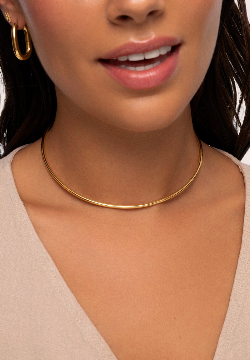 Goldfarbener Metall-Choker-Halsreifen mit einem eleganten, glatten, runden Design, der auf einem nackten Schlüsselbein ruht, kombiniert mit passenden Creolen.