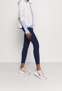 Hellrosa Zip-Jacke, blauer Langarmoberteil, marineblaue Leggings und hellrosa Sneakers. Merkmale sind glatte Texturen und ein sportliches Design.