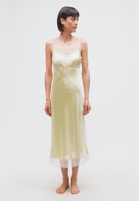 Γυναίκα που στέκεται ξυπόλυτη φορώντας ένα απαλό κίτρινο σατέν φόρεμα με δαντέλα, κοιτάζοντας προς τα δεξιά της σε λευκό απλό φόντο.