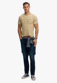 Camiseta de algodón beige con logo impreso, combinada con vaqueros azul oscuro y zapatillas blancas. Una camisa de cuadros azul marino anudada a la cintura.