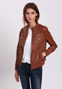 Giacca di pelle marrone con chiusura a zip, due tasche sul petto, polsini a coste e un design strutturato, abbinata a una camicia bianca e jeans blu.