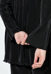 Schwarzes, plissiertes Oberteil mit langen Ärmeln, das aus einem strukturierten Stoff gefertigt ist und ausgestellte Bündchen hat. Hand greift den Stoff am Handgelenk zur Betonung.