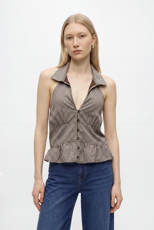 Ung kvinde iført brun- og hvidternet, ærmeløs knapper-top med peplumkant og blå jeans, stående mod hvid baggrund.