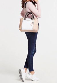 Sac à main beige et blanc avec une finition brillante, poignées noires, accents en chaîne argentée et une étiquette ; porté avec un jean noir et des baskets blanches.