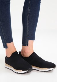 Svarta slip-on skor med en mjuk ovandel, vit sula och texturerad beige detalj. Bärs med blå jeans med fransad nederkant för kontrast.