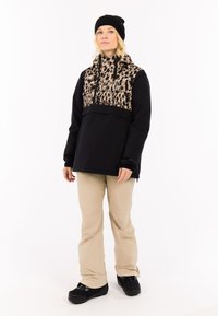 Schwarze Jacke mit Leopardenmuster im oberen Bereich, Fronttasche und hohem Kragen. Beigefarbene Hose und schwarze Schuhe runden das Outfit ab.