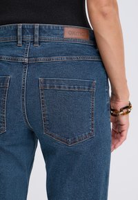 Jean en denim avec une teinte bleu foncé, présentant une étiquette de marque en cuir à l'arrière et des coutures visibles sur les poches. Texture lisse et coupe ajustée.