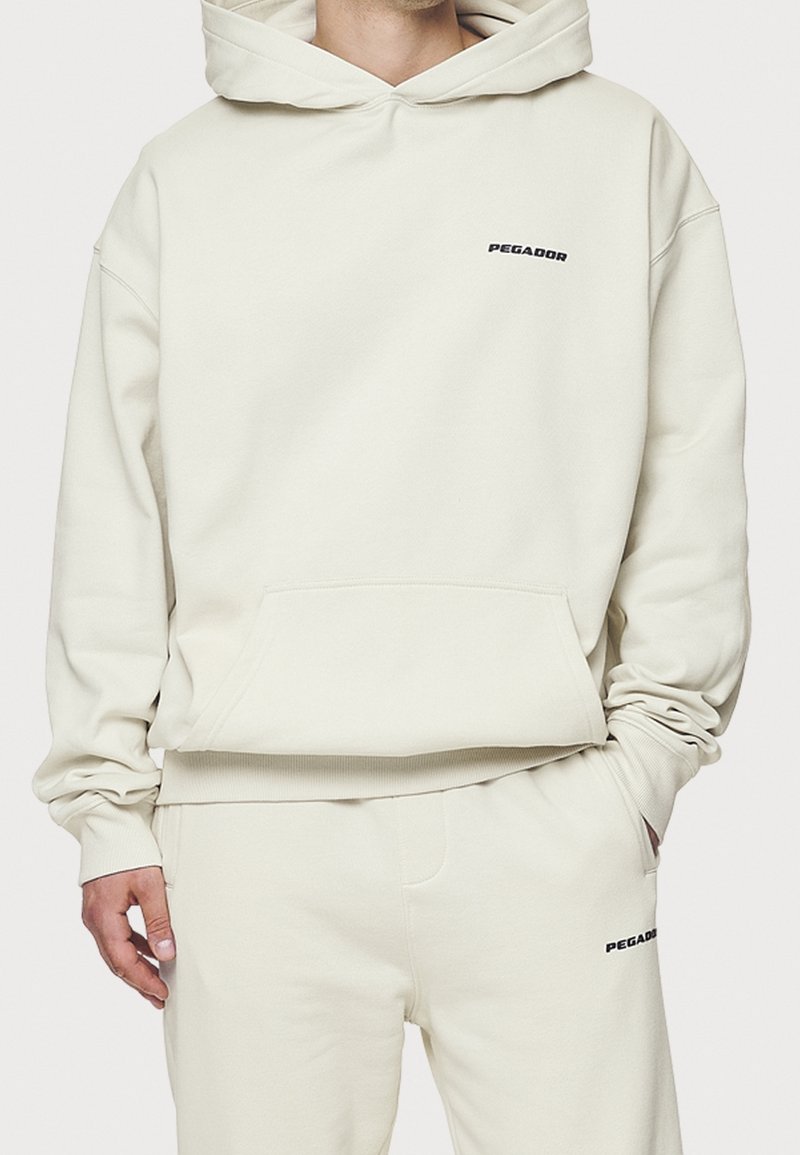 Modello che indossa un felpa di un leggero colore crema e pantaloni abbinati con il logo "PEGADOR" in nero. L'outfit presenta una vestibilità ampia e un cappuccio.
