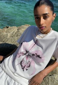 T-shirt en coton blanc avec un grand graphisme d'orchidée rose, encolure ronde et manches courtes, associé à une jupe blanche texturée.