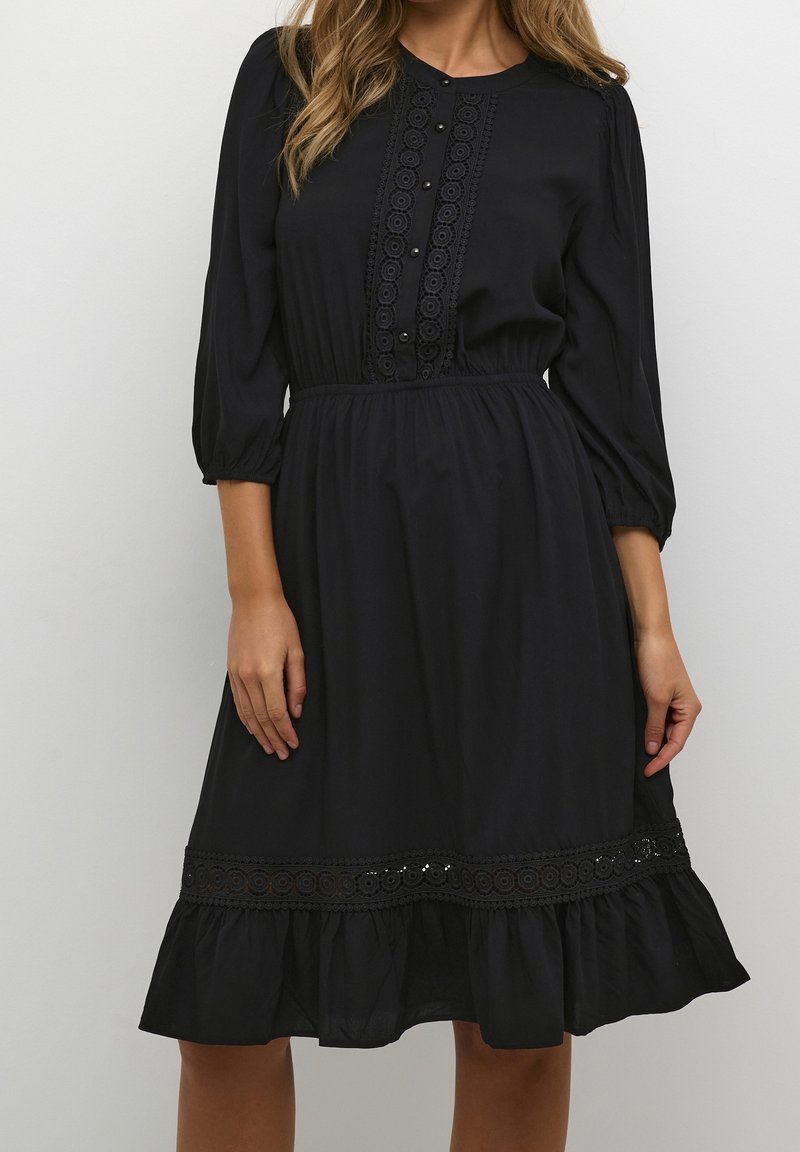 Femme portant une robe noire longueur genoux avec manches trois-quarts, dentelle décorative et boutons sur le devant et l'ourlet.