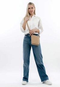 Beige läder crossbody-väska med en texturerad yta, rektangulär form och justerbar rem, uppvisad med en modell som bär en vit skjorta och blå jeans.