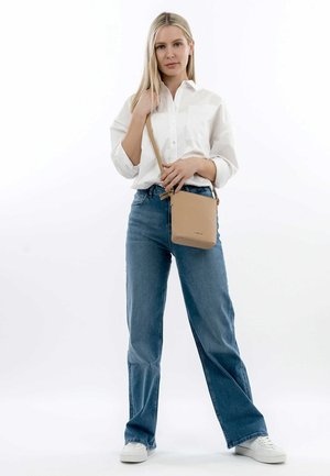 Borsa a tracolla in pelle beige con finitura strutturata, forma rettangolare e tracolla regolabile, mostrata con un modello che indossa una camicia bianca e jeans blu.