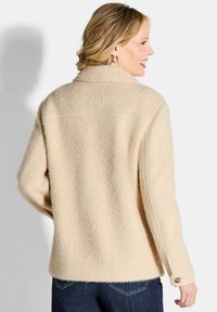 Beige wolmix trui met een relaxed fit, sjaalkraag, verlaagde schouders en knoopdetail op de manchetten. Zichtbare textuur en stiksels.