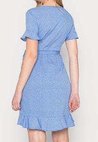 Robe bleue à pois blancs avec manches courtes à volants, taille cintrée par une ceinture et ourlet évasé en bas, en tissu léger.