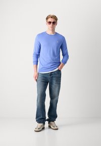 Helles blaues langärmliges Strickpullover mit gerippten Bündchen, kombiniert mit ausgewaschenen blauen Jeans und beigen Sneakers. Sonnenbrille mit hellrosa Gläsern.