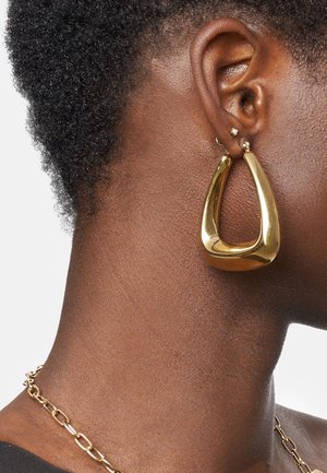 Boucles d'oreilles en or en forme de cerceau avec un design allongé et courbé. Texture lisse et brillante avec une esthétique moderne et minimaliste. Portées sur un lobe d'oreille.