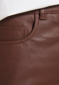 Détail d'un vêtement en cuir marron montrant les coutures de la poche et de la passants de ceinture avec un tissu blanc partiellement visible au-dessus.