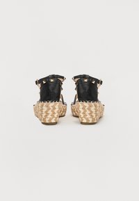 Sandales compensées en cuir noir avec des accents cloutés et une semelle en jute tressé. La vue de dos met en avant les sangles et la surface en cuir texturé.
