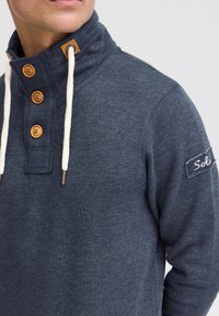 Suéter de color azul marino con un cuello alto, botones de madera y cordones blancos. Presenta una etiqueta de tela con la inscripción "Sol." Textura suave.