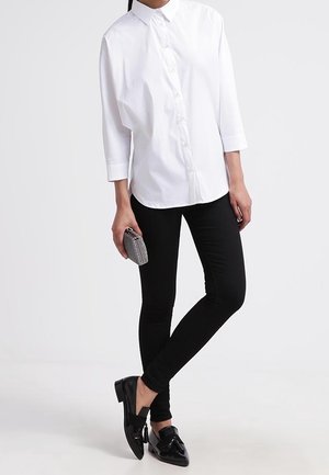 Femme portant une chemise blanche à boutons, un pantalon slim noir, des mocassins noirs, tenant une petite pochette argentée sur un fond uni.