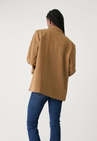 Veste marron oversize en tissu doux, avec une coupe décontractée, des manches larges et un empiècement unique dans le dos. Portée avec un jean bleu.
