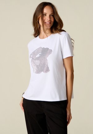 Donna con capelli castani lunghi che indossa una maglietta bianca con un motivo astratto a pois e pantaloni neri, sorridente e guarda a destra.