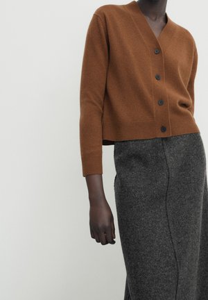 Model draagt een bruine cardigan met knopen, lange mouwen en een grijze wollen rok met hoge taille, voor een effen achtergrond.