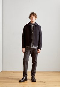 Giacca nera con zip e colletto, camicia grigia con bottoni sotto, pantaloni grigi a fantasia e scarpe nere. Tessuto liscio, vestibilità su misura.