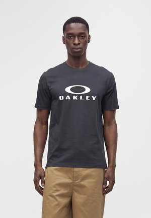 Mężczyzna stojący przodem, ubrany w czarną koszulkę z białym logo Oakley i beżowe spodnie na jednolitym jasnym tle.