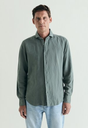 Hombre con el cabello corto y oscuro, vistiendo una camisa verde de manga larga abotonada y vaqueros azul claro, de pie frente a un fondo claro y liso.