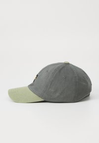 Căciulă de baseball gri cu o vizieră verde deschis. Prezintă o pată neagră cu un design galben pe partea din față, realizată din bumbac cu o textură netedă.