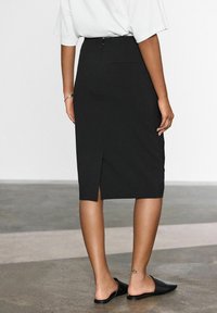 Jupe crayon noire avec une fente à l'arrière, taille haute et fermeture éclair dissimulée. Portée avec des chaussures noires à enfiler, jambes visibles avec un tatouage en forme d'étoile.
