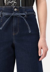 Calças de ganga escuras de cintura alta com um cinto atado, apresentando um fecho de botão e bolsos frontais. Textura suave com pormenores de costura subtis.