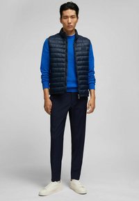 Marineblaues Puffer-Westen über einem blauen Pullover, kombiniert mit dunklen Hosen und weißen Sneakers. Verfügt über einen Reißverschluss vorne und gerippte Bündchen.