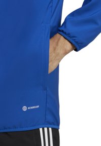 Blaue Sportjacke mit glatter Textur, verfügt über eine Seitentasche und ein rundes Logo mit der Aufschrift "AEROREADY" nahe dem Saum. Elastische Bündchen.