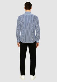 Camicia a righe blu e bianche con maniche lunghe e colletto con bottoni, abbinata a pantaloni neri e sneakers bianche. Sfondo semplice.