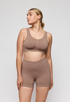 Vrouw draagt een naadloze nude sportbeha en een korte broek met een hoge taille in dezelfde kleur, staande met haar hoofd naar de zijkant gedraaid tegen een effen achtergrond.