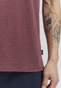 Burgunderfarbenes T-Shirt mit kurzen Ärmeln aus strukturiertem Stoff; sichtbare Nähte und Markenlabel am Saum; getragen mit blauen Hosen.