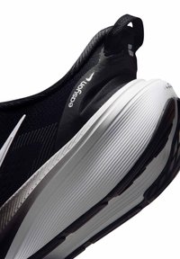 Μαύρο και άσπρο παπούτσι για τρέξιμο Nike Easyon με παχιά μαλακή σόλα και θηλιά στη φτέρνα, εμφανίζεται από γωνιακή κοντινή λήψη.