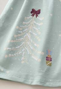 Chemise en coton vert clair ornée d'un sapin de Noël stylisé avec des sequins, un nœud bordeaux et de petites boîtes cadeaux de différentes couleurs et motifs.