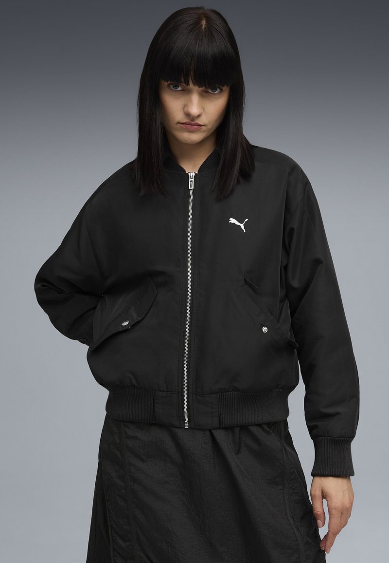 Veste bomber noire avec un zip sur le devant, des poignets et un ourlet côtelés. Comprend des poches latérales et un petit logo blanc sur la poitrine. Texture de tissu lisse.